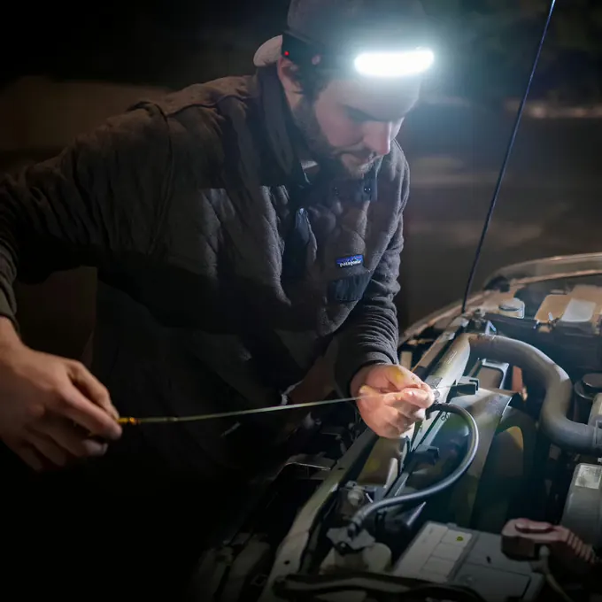 Mechaniker beugt sich über einen Motorraum, beide Hände an den Bauteilen, beleuchtet von einer Stirnlampe