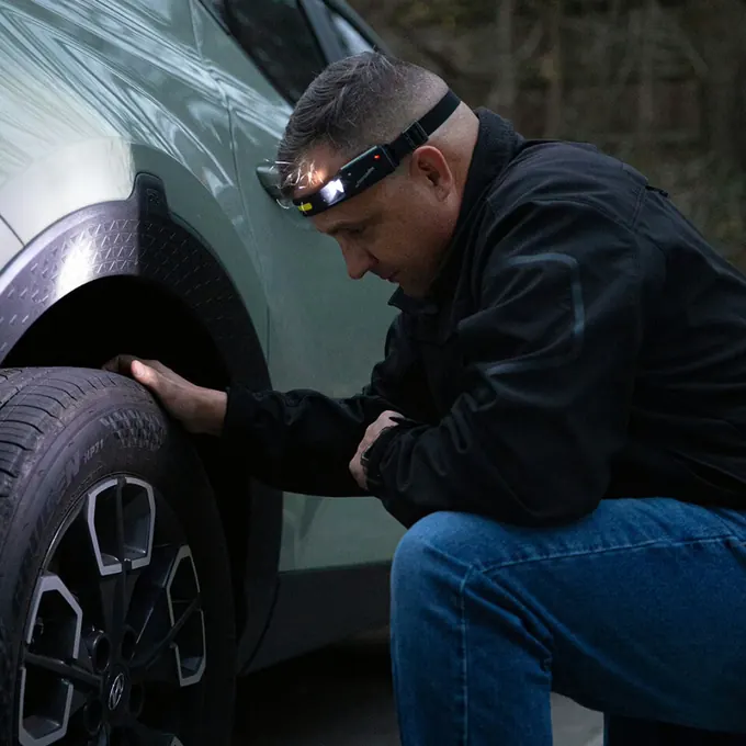Mann kniet nachts neben einem Auto und prüft einen Reifen im Licht seiner Stirnlampe