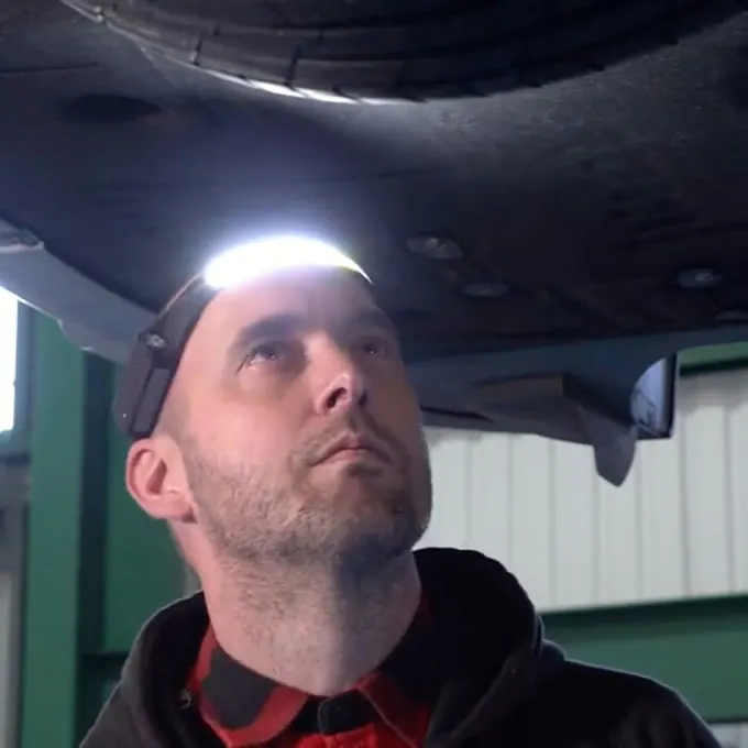 Mann blickt mit eingeschaltetem breitem Licht der Stirnlampe auf die Unterseite eines Fahrzeugs